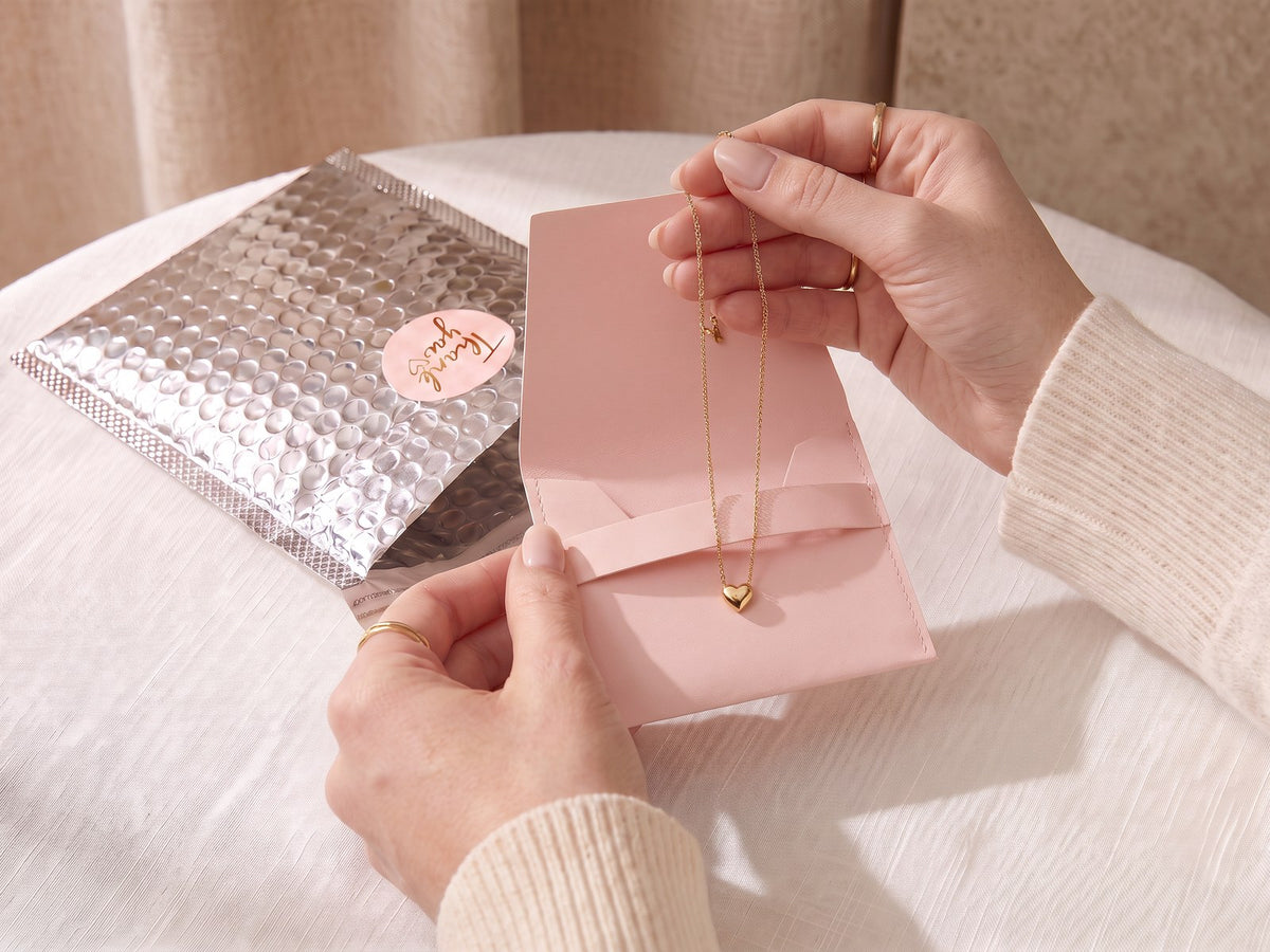 Collier cœur doré sorti de son écrin pochette rose, devant son enveloppe argentée Maison Fermoir